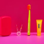 Colorful dental-care still life: yellow toothbrush, orange toothpaste tube, red case, and small dental tools on a pink surface.
