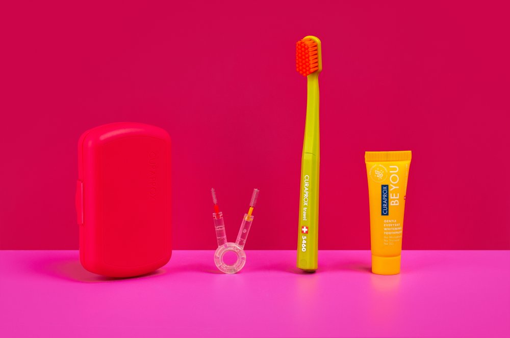 Colorful dental-care still life: yellow toothbrush, orange toothpaste tube, red case, and small dental tools on a pink surface.