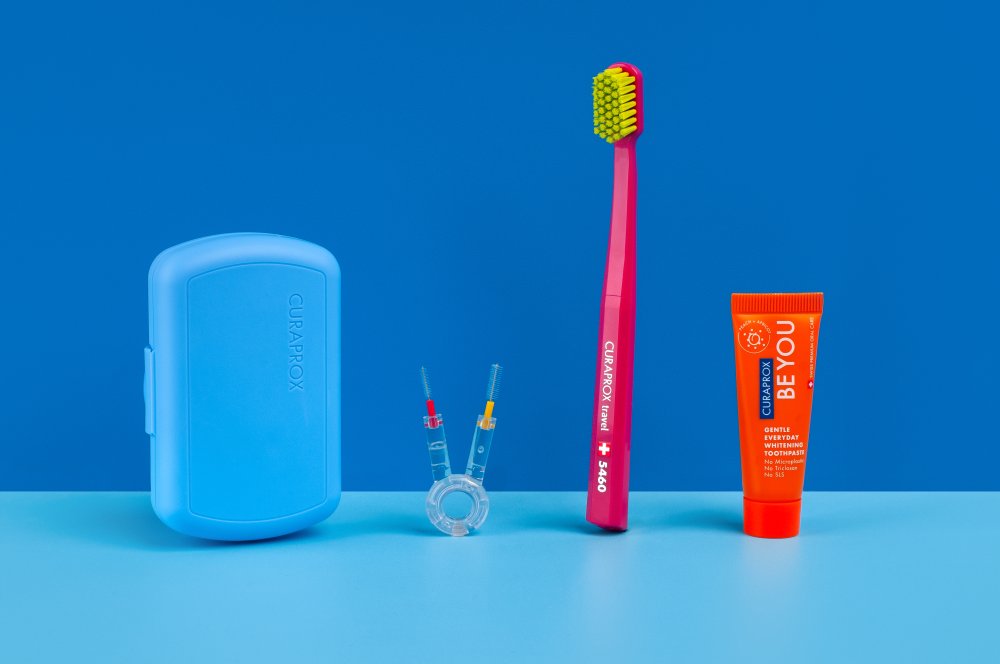 Curaprox Travel Set Modrá 4 Blue travel dental kit with a curved case, pink toothbrush, orange Be You toothpaste, and interdental brushes on a blue surface.