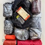 Stacked hiking gear on a light wood floor: black center backpack, gray and maroon Hyperlite Mountain Gear bags, and red/orange stuff sacks with a water bottle.
