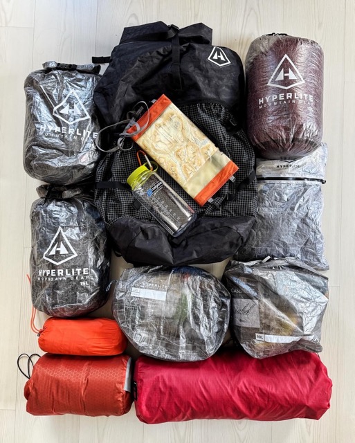 Stacked hiking gear on a light wood floor: black center backpack, gray and maroon Hyperlite Mountain Gear bags, and red/orange stuff sacks with a water bottle.