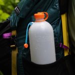 Hydration bottle with orange cap tucked in a backpack side pocket, droplets on the bottle in a wet outdoor setting.