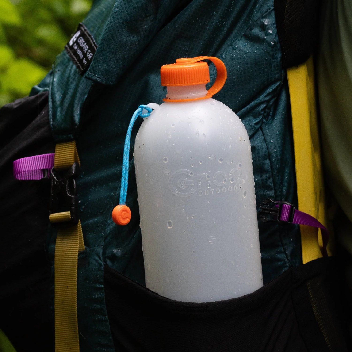 Hydration bottle with orange cap tucked in a backpack side pocket, droplets on the bottle in a wet outdoor setting.