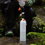 Hiker filling a hydration reservoir from a bottle outdoors on a damp rock, surrounded by ferns and moss.