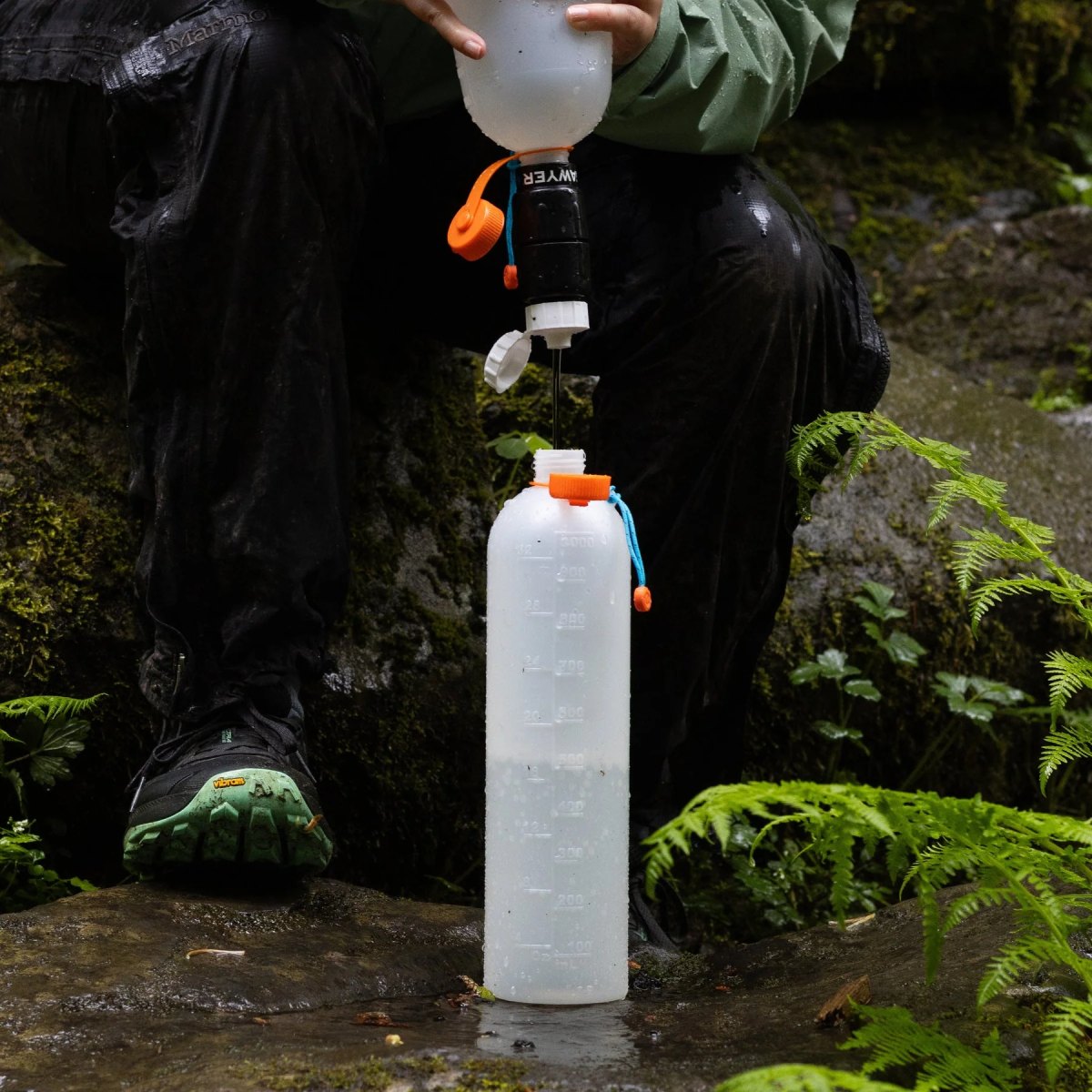 Hiker filling a hydration reservoir from a bottle outdoors on a damp rock, surrounded by ferns and moss.