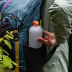 Side pocket hydration bottle on a hiker wearing a teal rain jacket, with droplets on fabric and a yellow strap nearby.