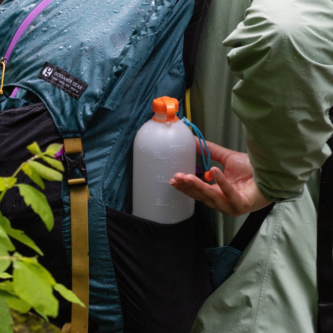 Side pocket hydration bottle on a hiker wearing a teal rain jacket, with droplets on fabric and a yellow strap nearby.