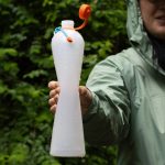 Person in a green rain jacket holding a white translucent squeeze bottle with an orange cap outdoors.