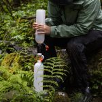 Person outdoors assembling a portable water filter system between two plastic bottles in a mossy forest setting, wearing a green rain jacket.