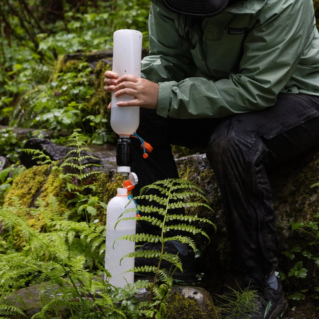 Person outdoors assembling a portable water filter system between two plastic bottles in a mossy forest setting, wearing a green rain jacket.