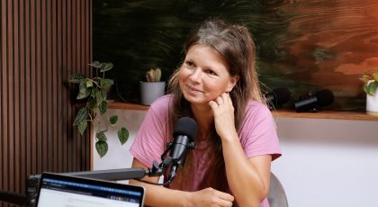 Woman in a pink shirt sits at a podcast studio desk, smiling with a hand resting on her chin, two microphones in front of her, and a laptop nearby.