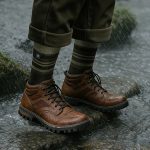 Brown leather hiking boots with dark striped socks standing on wet rocks in the rain.