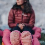Person wearing a red jacket sits outdoors on an orange sleeping pad, pink boots facing the camera.