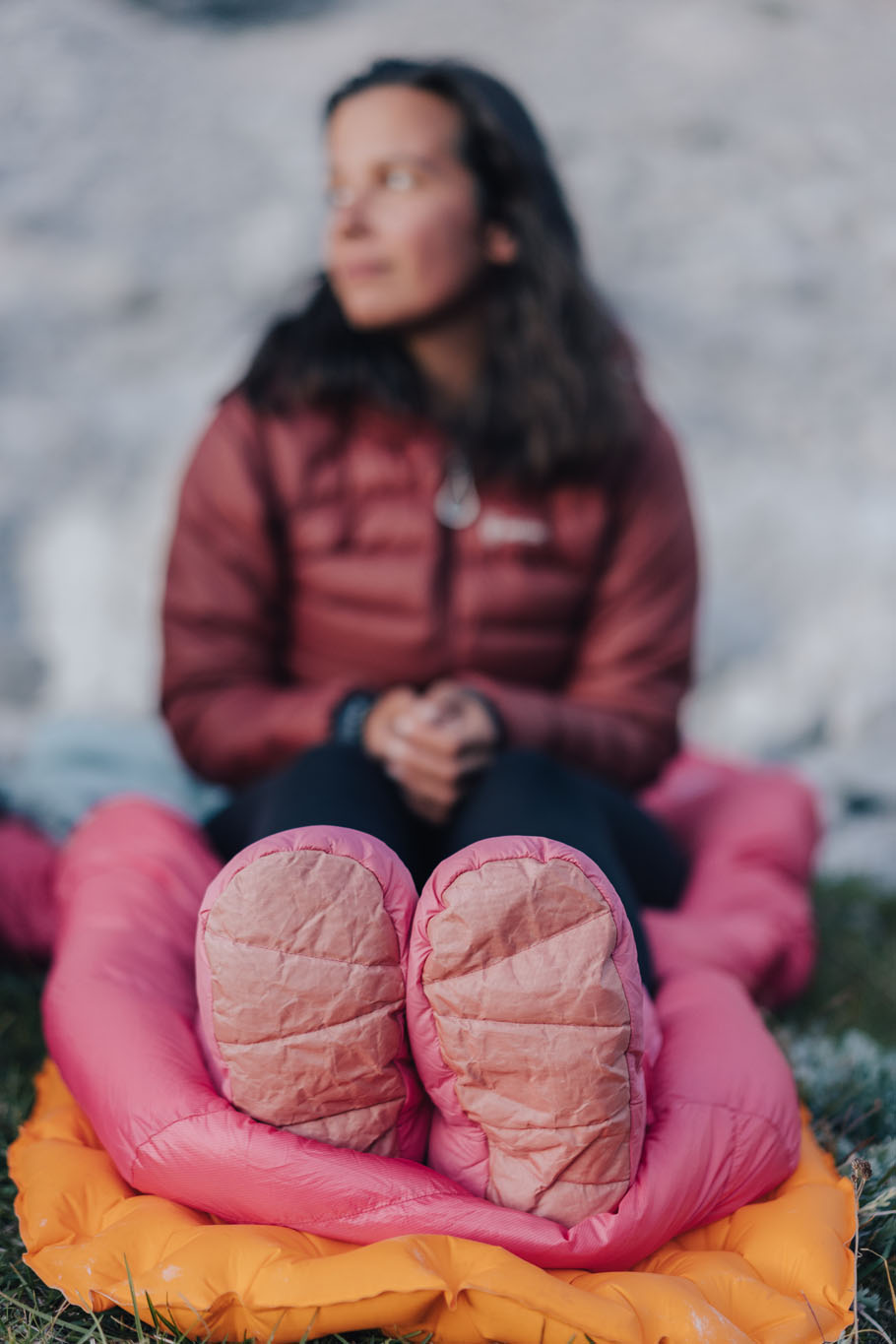 Person wearing a red jacket sits outdoors on an orange sleeping pad, pink boots facing the camera.