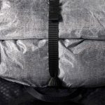 Close-up of a gray backpack with a vertical black strap and buckle on textured fabric.
