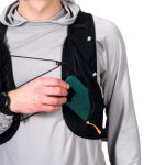 Person wearing a black running vest places a green speckled smartphone into a front pocket while wearing a gray long-sleeve top and a watch.