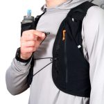Runner wearing a black hydration vest with a blue-topped water bottle in a front pocket, holding the bite valve to drink during a run.