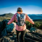 Hiker on rocky mountaintop with arms outstretched, wearing pink jacket and gray backpack against blue sky.