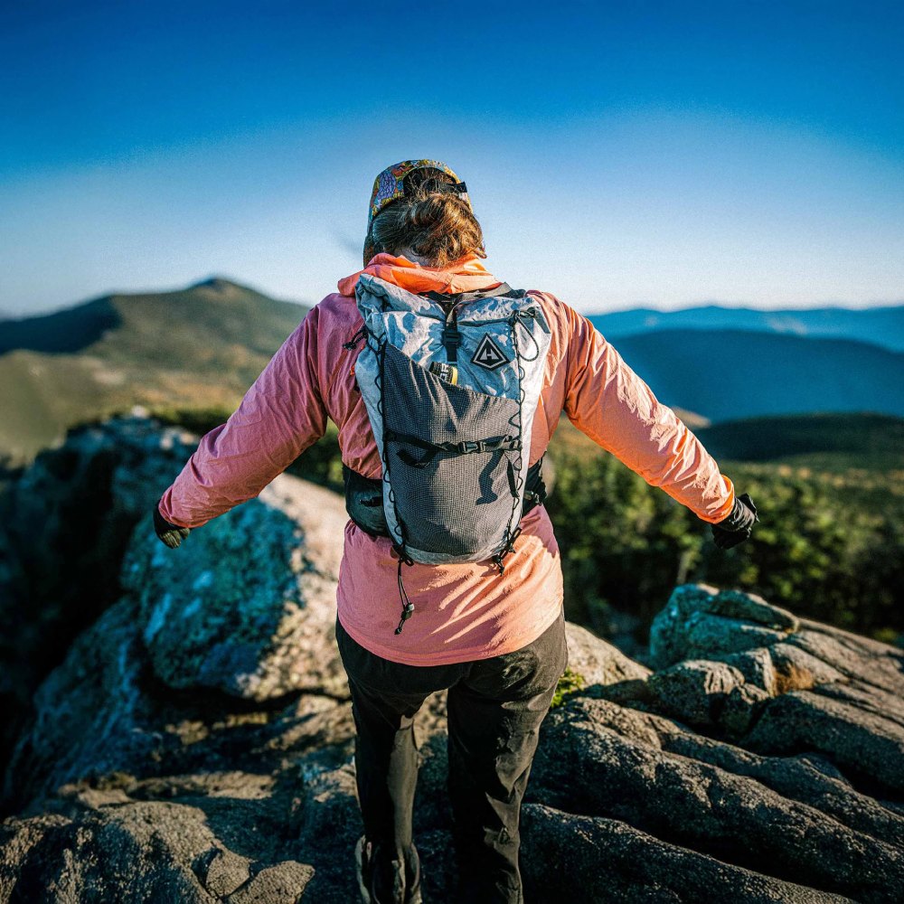 Hiker on rocky mountaintop with arms outstretched, wearing pink jacket and gray backpack against blue sky.