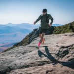 Man trail-running on rocky mountain ridge with a backpack, wearing a cap, green top, black shorts, and bright pink socks against a blue sky and distant mountains.