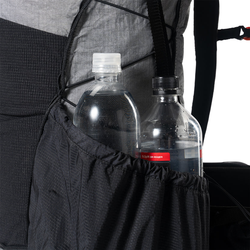 Backpack with two water bottles in the side pocket, one clear bottle with a white cap and the other with a black cap.