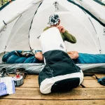 Person kneeling inside a white tent on a wooden deck, adjusting a sleeping bag as camping gear and a propane stove sit nearby.
