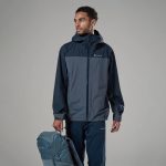 Montane Mens Norste Lite Jacket Eclipse Blue 02 Man wearing a two-tone navy Montane rain jacket with hood, standing against a neutral backdrop, holding a matching backpack at the bottom left.