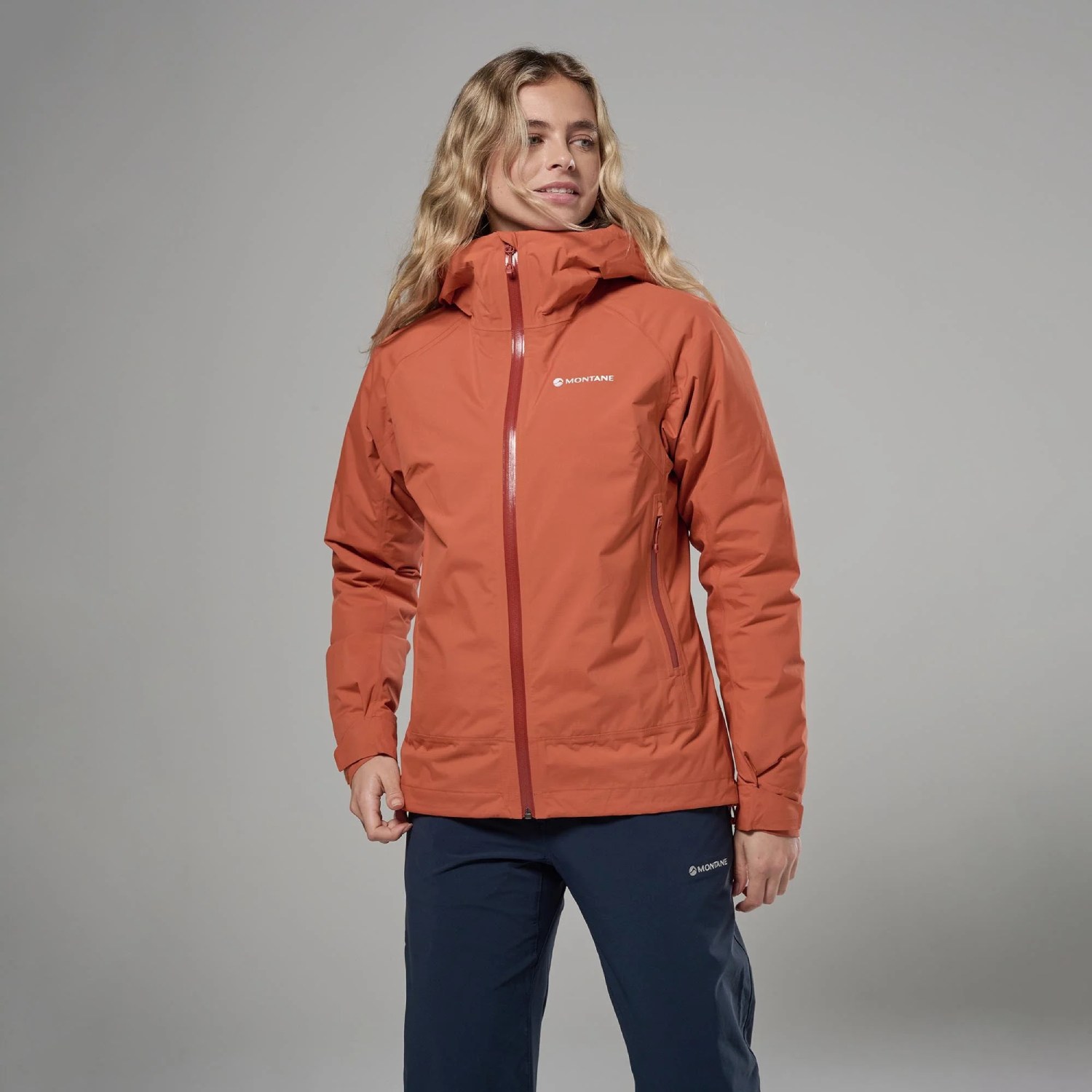Montane Womens Norste Lite Jacket Lava 01 1 Woman wearing an orange Montane lightweight jacket with a high collar, zipped front, and navy pants against a neutral backdrop.