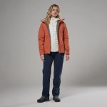 Montane Womens Norste Lite Jacket Lava 03 Woman in coral waterproof jacket over a white quilted vest, navy hiking pants, and dark hiking shoes, standing against a gray studio backdrop.