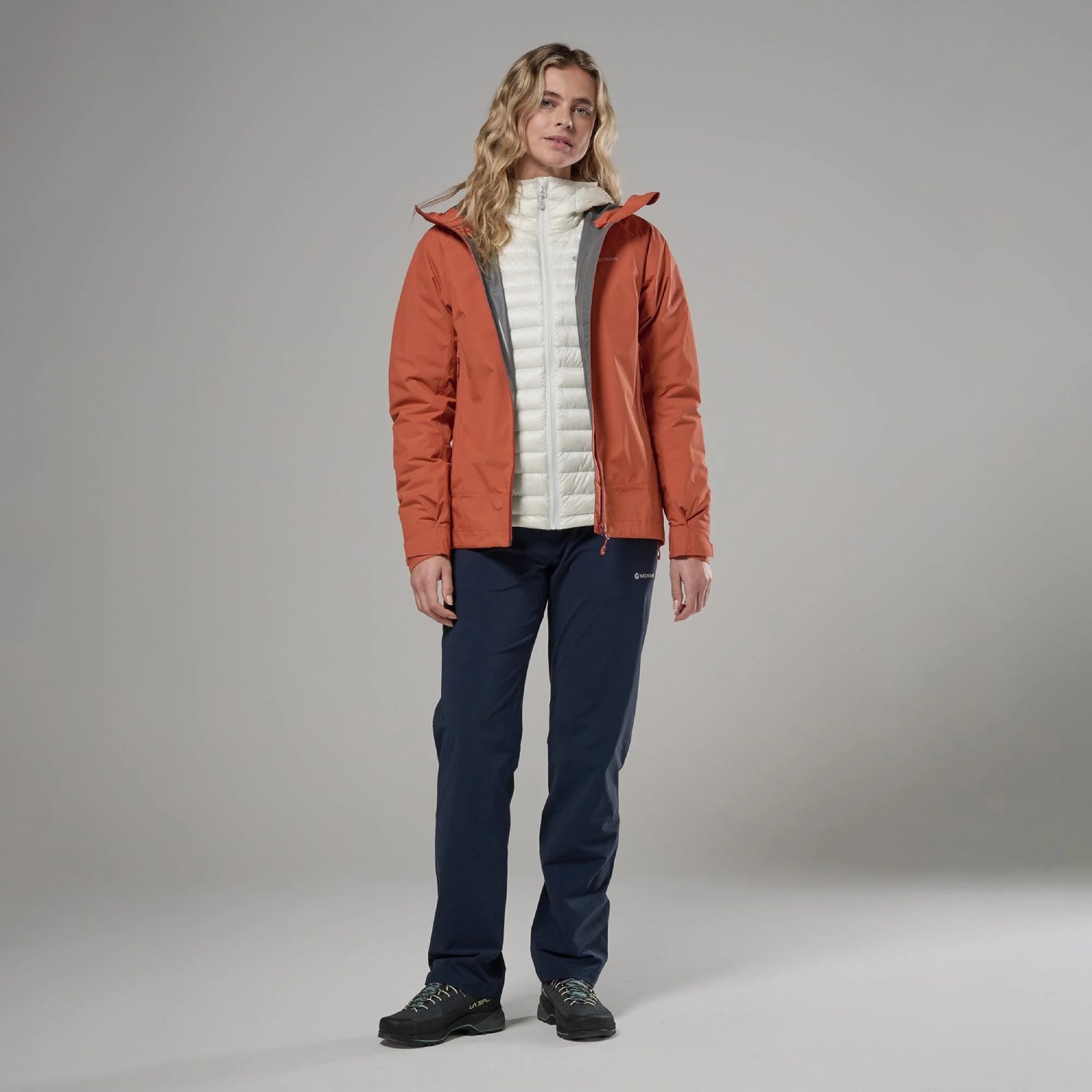 Montane Womens Norste Lite Jacket Lava 03 Woman in coral waterproof jacket over a white quilted vest, navy hiking pants, and dark hiking shoes, standing against a gray studio backdrop.