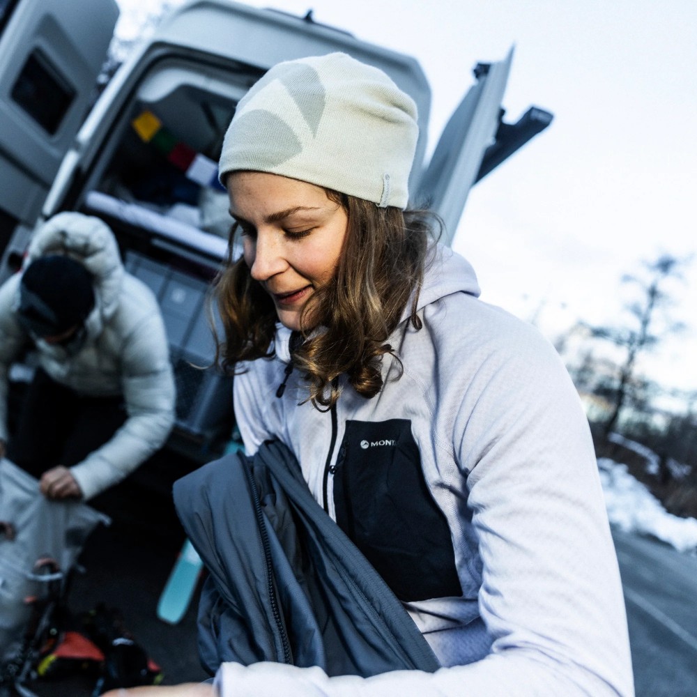 Montane Womens Zepher Hoodie Platinum 09 Woman in a light gray outdoor jacket and white beanie packs a dark jacket by an open van on a chilly, snowy day.